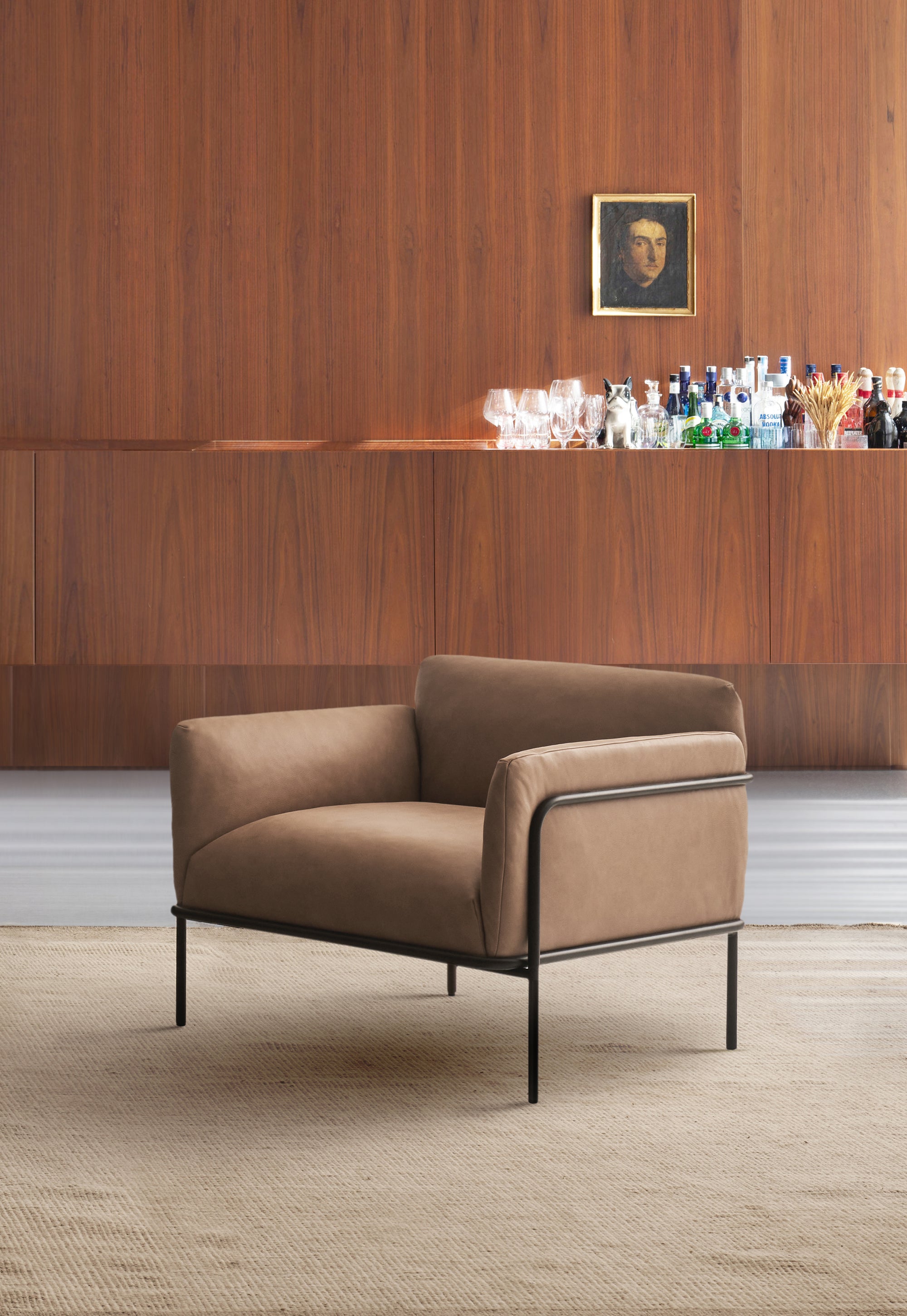The Scribe Lounge Chair | Leather by Wooden Stories, featuring a modern beige design with a Bauhaus-inspired metal frame and black legs, sits on a textured rug before a wooden wall. A bar area behind displays bottles, glasses, and a small framed portrait.