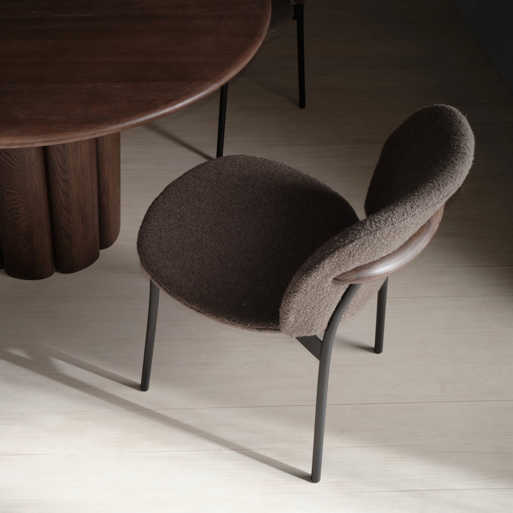 The Wooden Stories Solace Dining Chair | Fabric features a curved, textured brown seat and backrest, premium steel legs, and a rounded wooden accent, positioned beside a dark wood table on a light floor.
