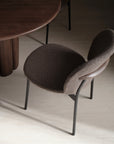 The Wooden Stories Solace Dining Chair | Fabric features a curved, textured brown seat and backrest, premium steel legs, and a rounded wooden accent, positioned beside a dark wood table on a light floor.