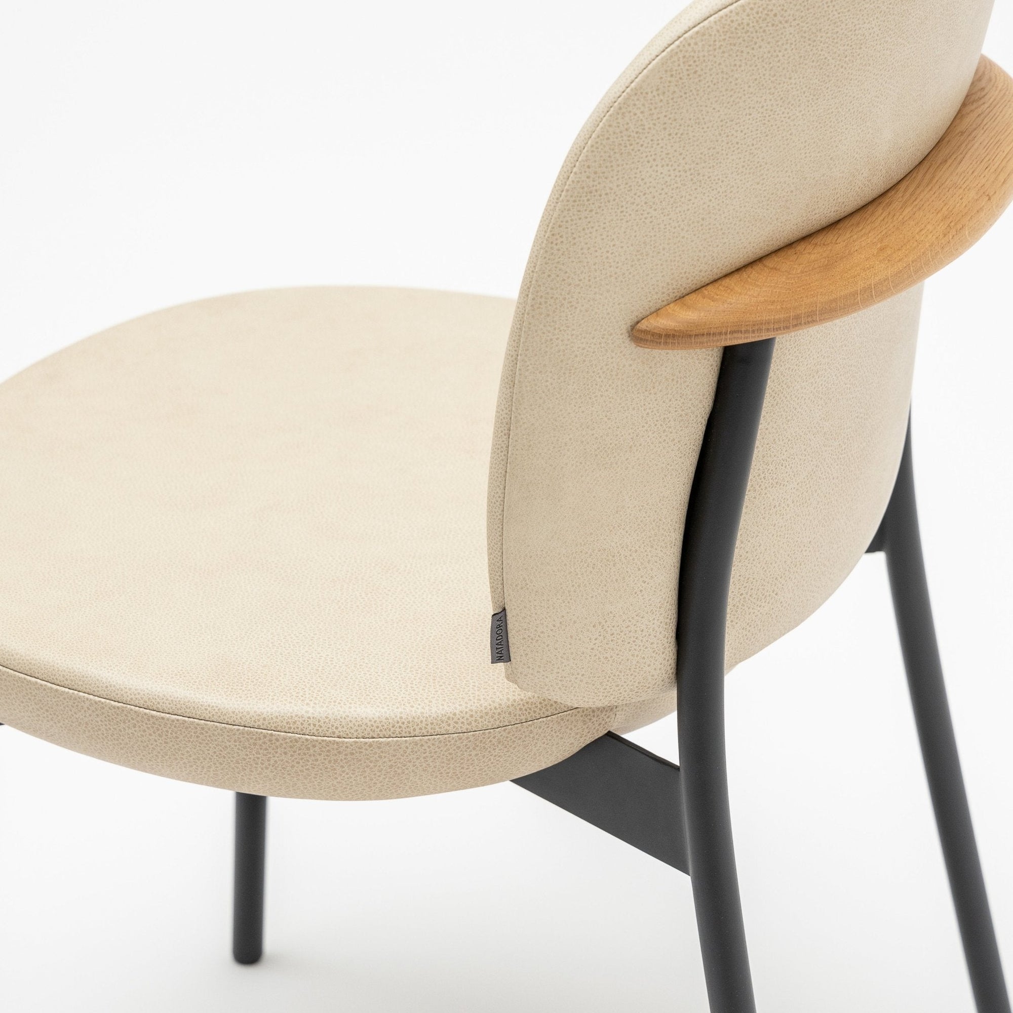 A close-up of the Solace Dining Chair | Leather by Wooden Stories, featuring a beige upholstered seat and backrest, black steel legs, and a curved wooden accent on the backrest, set against a plain white background.