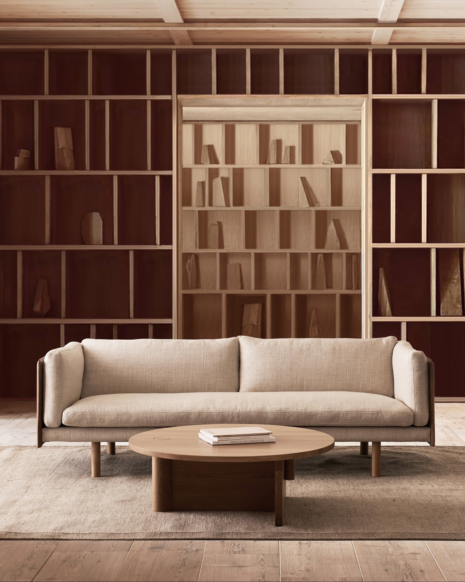 A minimalist living room with the Sonder Sofa by Wooden Stories—featuring high-density foam cushions, fabric upholstery with a leather back—in beige, complemented by a round wooden coffee table and built-in wooden bookshelves holding a few scattered books, all in neutral tones.