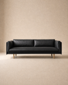 Centered against a plain beige background, the Sonder Sofa | Leather by Wooden Stories features modern Danish lines with a slim profile and wooden legs. Its black leather upholstery is complemented by feather-filled cushions, adding extra comfort to its contemporary design.