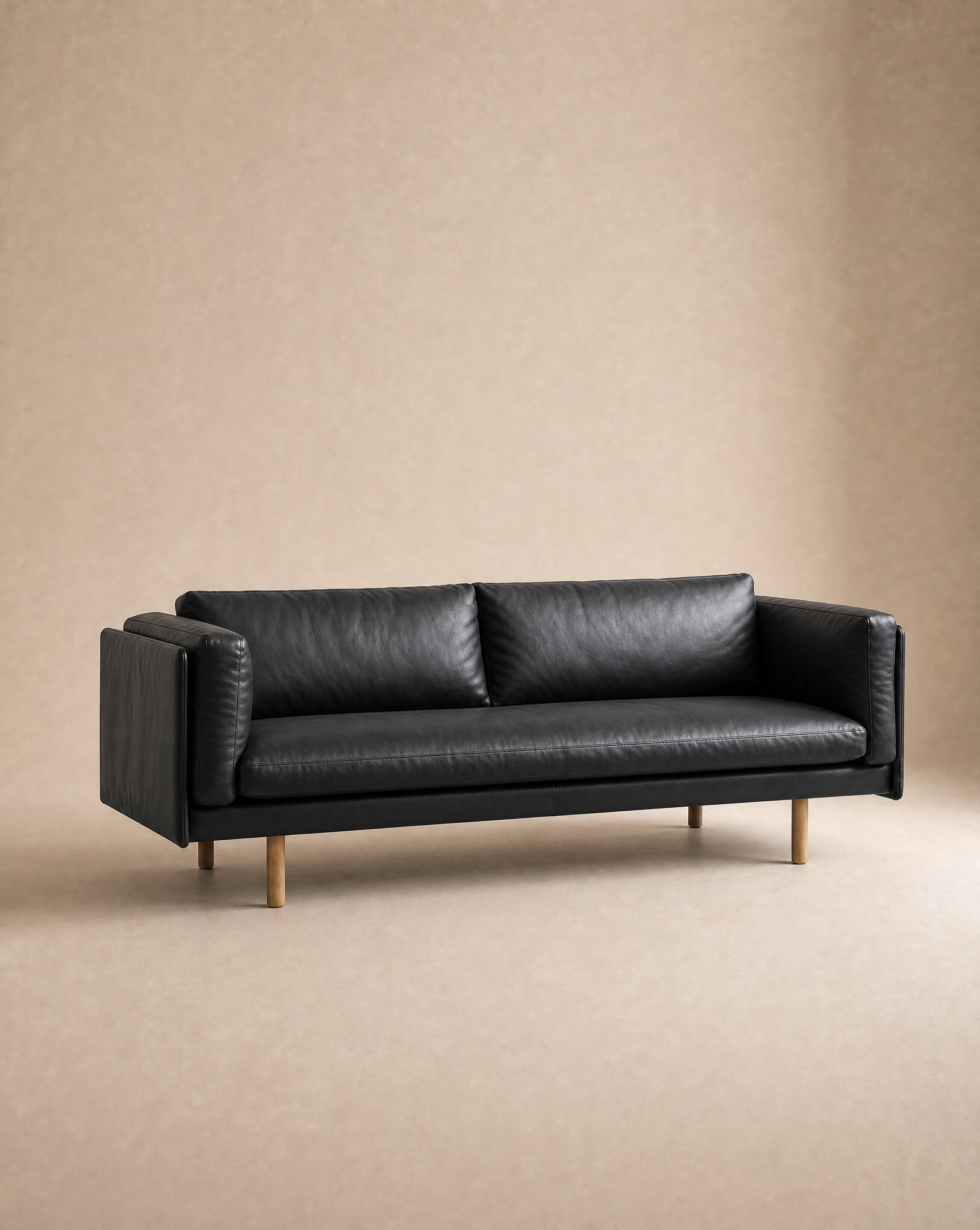 The Sonder Sofa | Leather by Wooden Stories—a modern black leather piece with Danish lines, slim proportions, and wooden legs—is set in an empty room with beige walls and floor.
