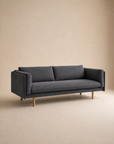 The Sonder Sofa | Fabric by Wooden Stories features a modern, minimalist design in dark gray, with two feather-filled cushions and wooden legs. Its clean lines, linear frame, and straight armrests create a sleek look, making it stand out against the neutral beige background.
