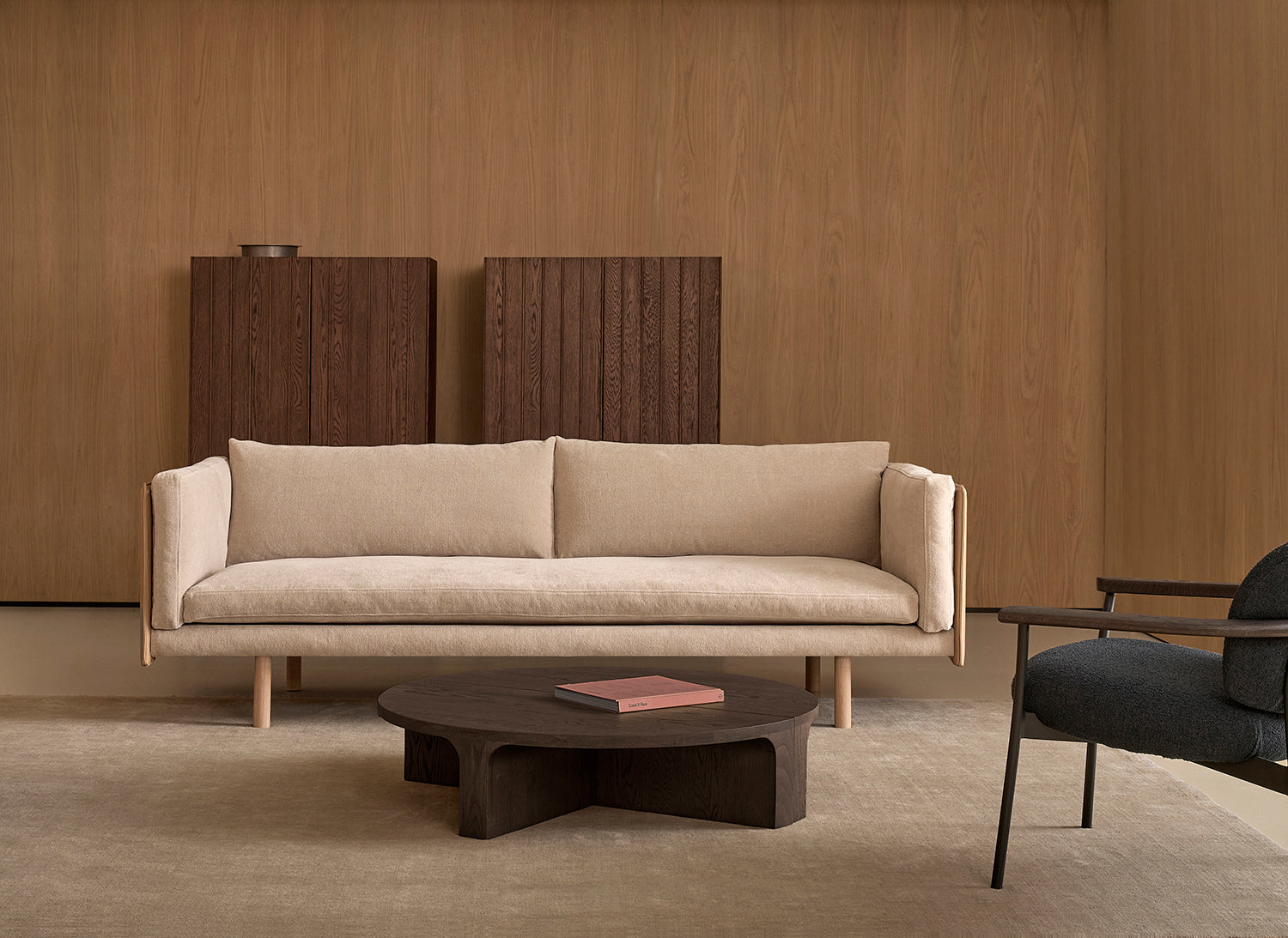 A minimalist living room with a Wooden Stories Sonder Sofa | Fabric with Leather Back in beige, featuring high-density foam cushions, a dark wood coffee table holding a book, two tall wooden cabinets, wooden wall paneling, and part of a dark armchair.