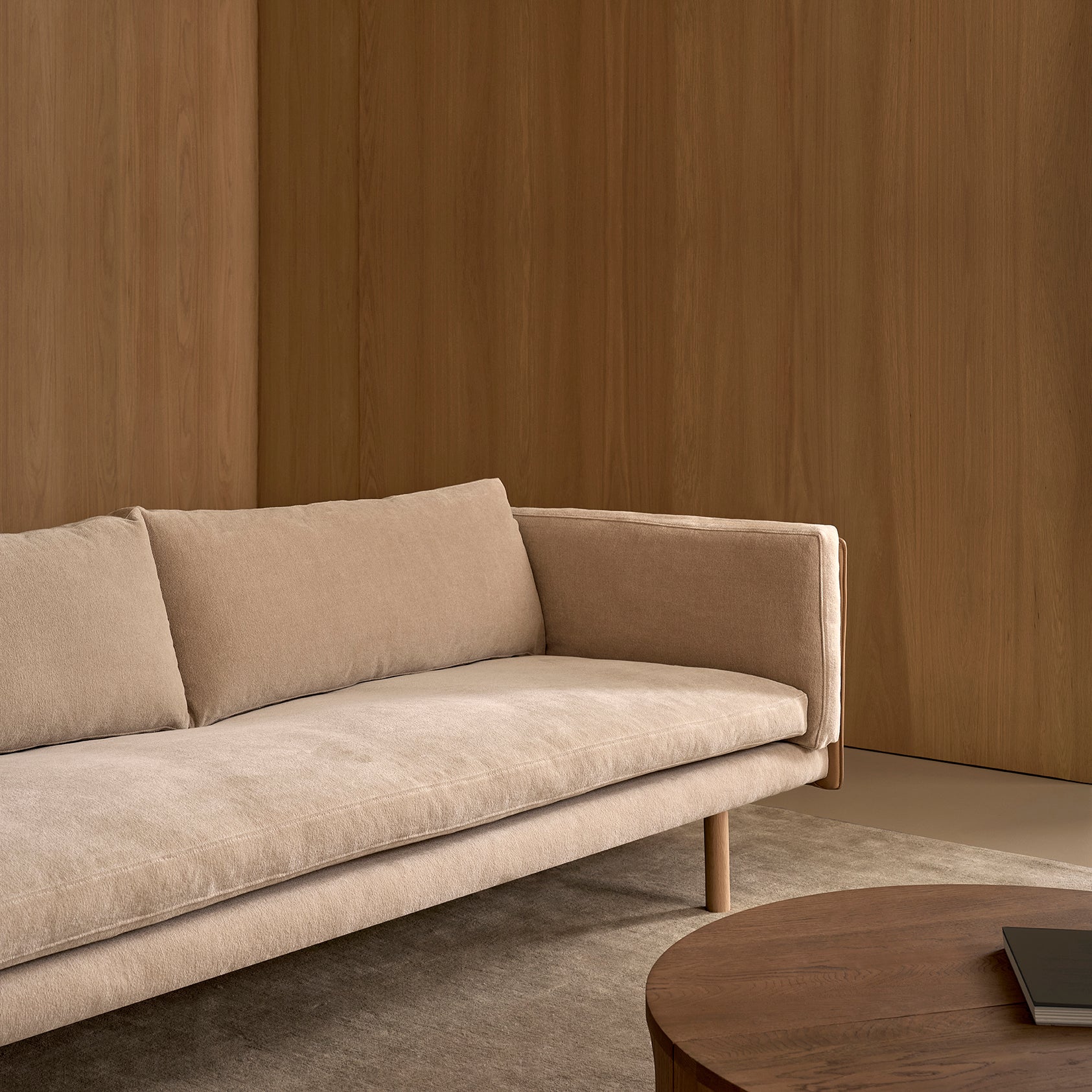A Sonder Sofa | Fabric with Leather Back by Wooden Stories, featuring high-density foam cushions and wooden legs in a beige finish, sits on a gray rug beside a round wooden coffee table. Smooth wooden panel walls in the background create a warm, minimalist atmosphere.
