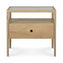 The Spindle Nightstand - Oak by Ethnicraft features solid oak construction, a glass top, an open shelf below, and a drawer with a central round cut-out handle.