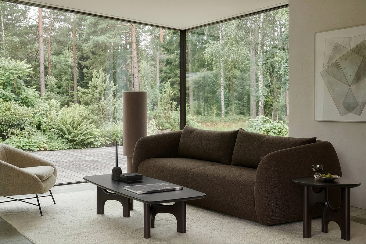 Modern living room featuring the Ethnicraft Figur Sofa | 3 Seater in brown, accompanied by a matching armchair, coffee table, and side table. Abstract wall art adorns the space, while expansive floor-to-ceiling windows reveal a lush forest view outside. Neutral tones and minimalist decor enhance the cozy, serene atmosphere—ideal for contemporary living.
