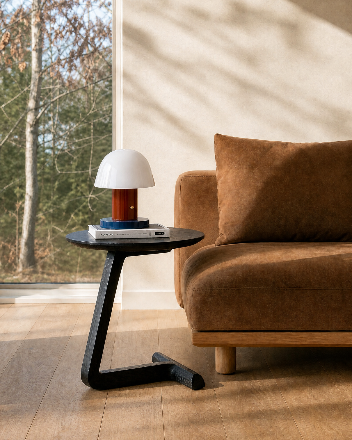 A modern living room features a brown suede sofa and the SOBU Teena Side Table in black ash, its cantilevered top displaying a small white and red lamp, books, and sunlight pouring through large windows with trees outside.