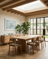 A modern dining room with the SOBU Tommaso Dining Table - Wood Legs and matching chairs, large windows, indoor plants, a sideboard of books, and a framed swimmer photo. Sunlight streams through skylights and glass doors to the garden.