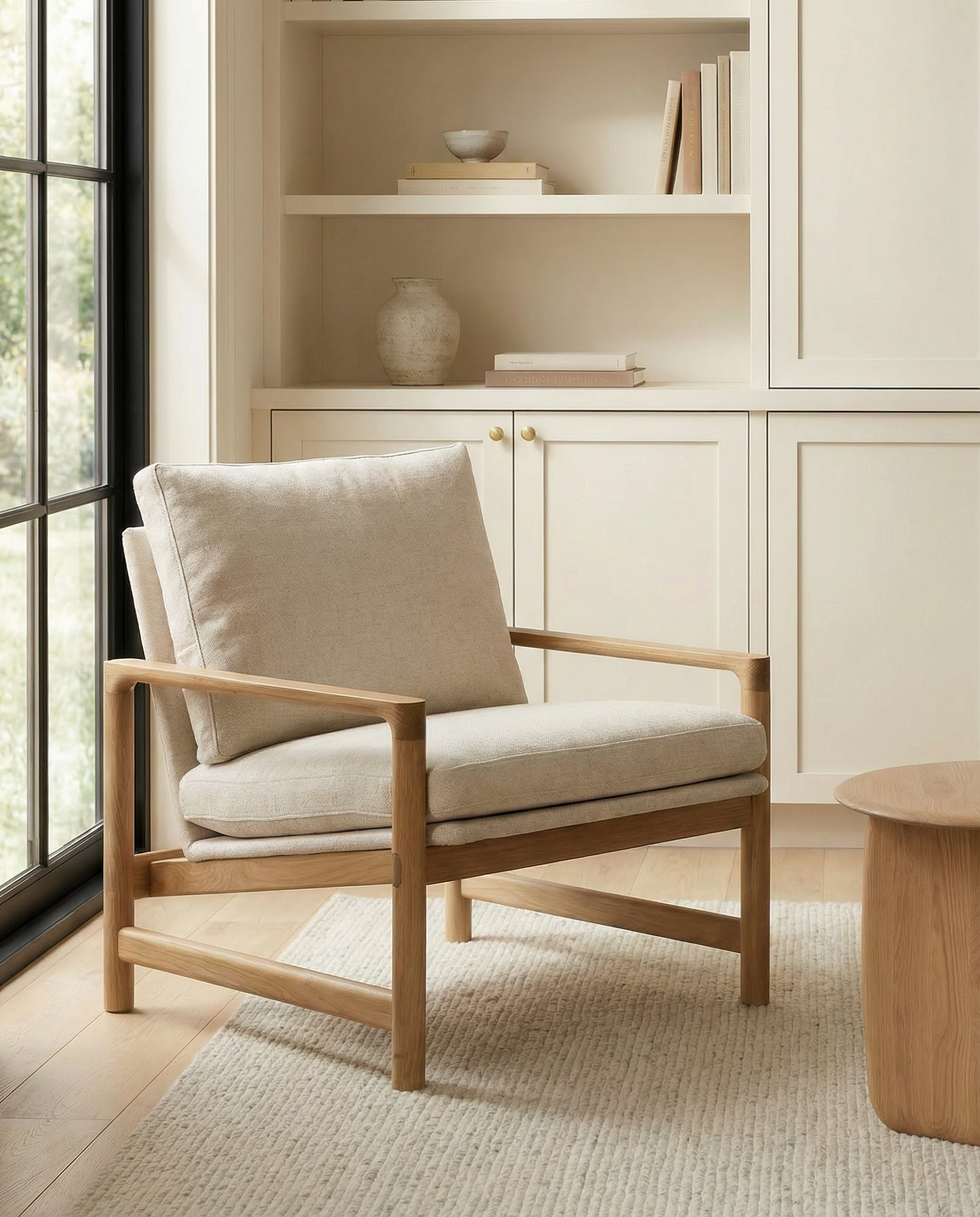 The Troubadour Lounge Chair by Wooden Stories, featuring a solid oak frame and beige fabric cushions, stands on a textured rug in a bright, minimalist room with built-in shelves, a round wooden table, and large windows inspired by Danish design.