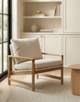The Troubadour Lounge Chair by Wooden Stories, featuring a solid oak frame and beige fabric cushions, stands on a textured rug in a bright, minimalist room with built-in shelves, a round wooden table, and large windows inspired by Danish design.