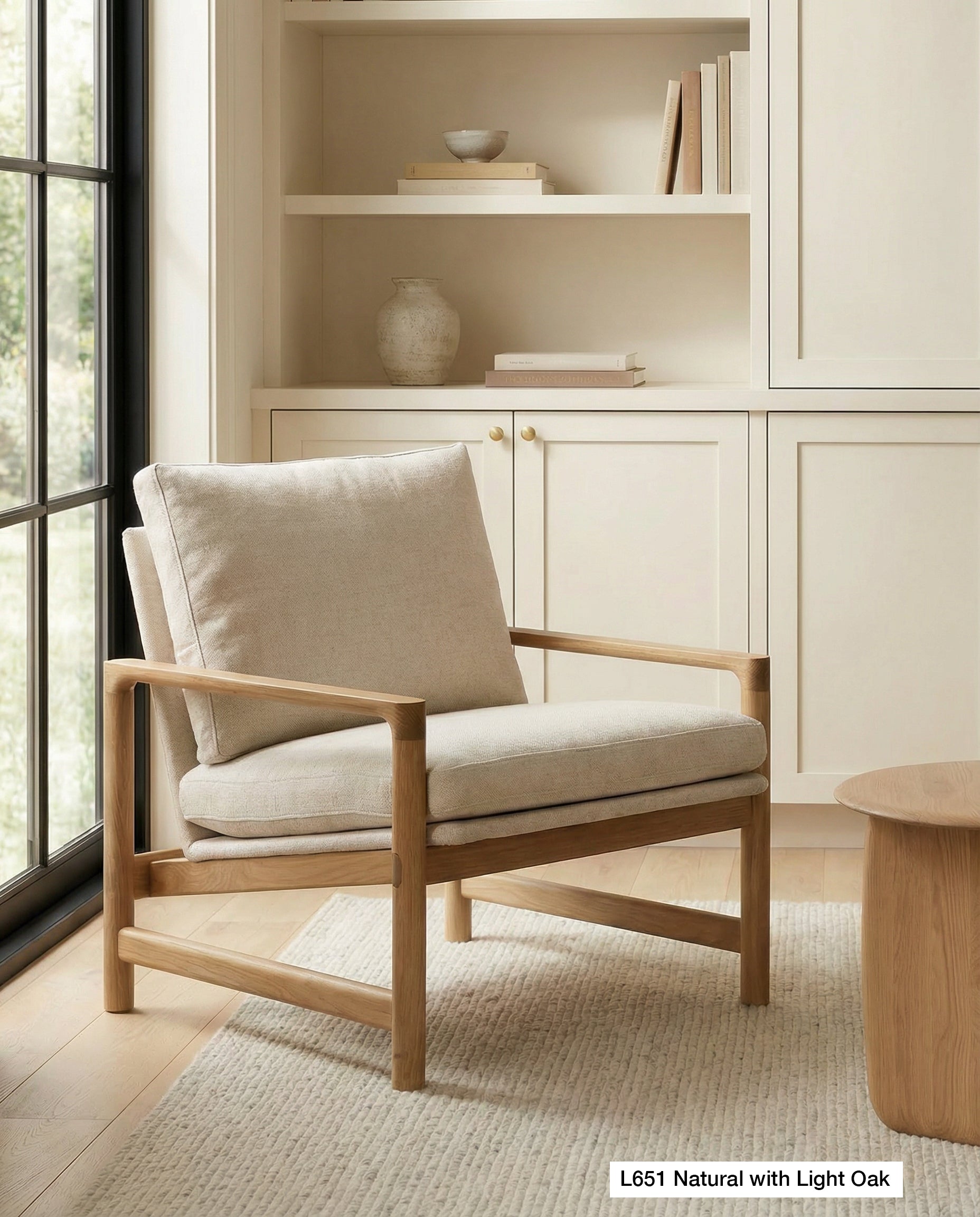 The Wooden Stories Troubadour Lounge Chair | Fabric, featuring a solid oak frame and beige cushions, is placed on a textured rug next to a wooden table, positioned before built-in shelves filled with books and decor in a softly lit, neutral-toned room.