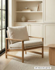 The Wooden Stories Troubadour Lounge Chair | Fabric, featuring a solid oak frame and beige cushions, is placed on a textured rug next to a wooden table, positioned before built-in shelves filled with books and decor in a softly lit, neutral-toned room.