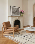 A cozy living room with the Wooden Stories Troubadour Lounge Chair in leather, featuring a solid oak frame, a white fireplace topped with a framed black-and-white photo and pink flowers, a modern coffee table, and a cream rug with a black grid pattern.