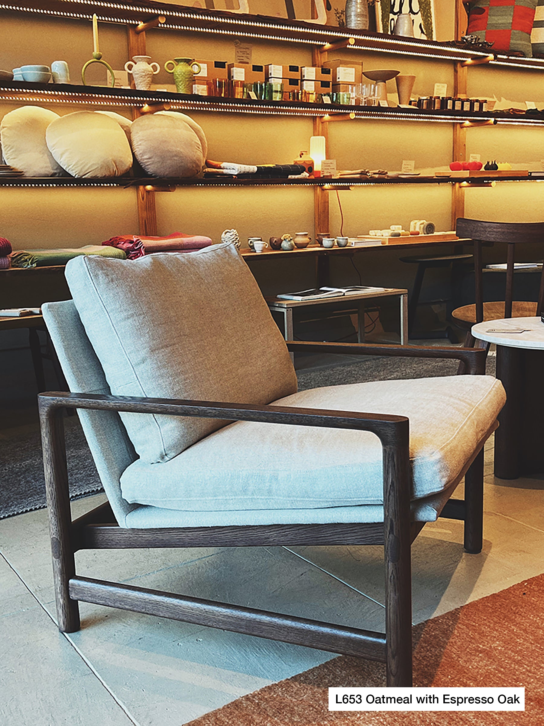 A Troubadour Lounge Chair | Fabric by Wooden Stories from the Danish Collection, with oatmeal-colored cushions and a solid oak frame in an espresso finish, sits in a cozy, well-lit store surrounded by shelves of home decor and ceramics. A small label reads, L653 Oatmeal with Espresso Oak.