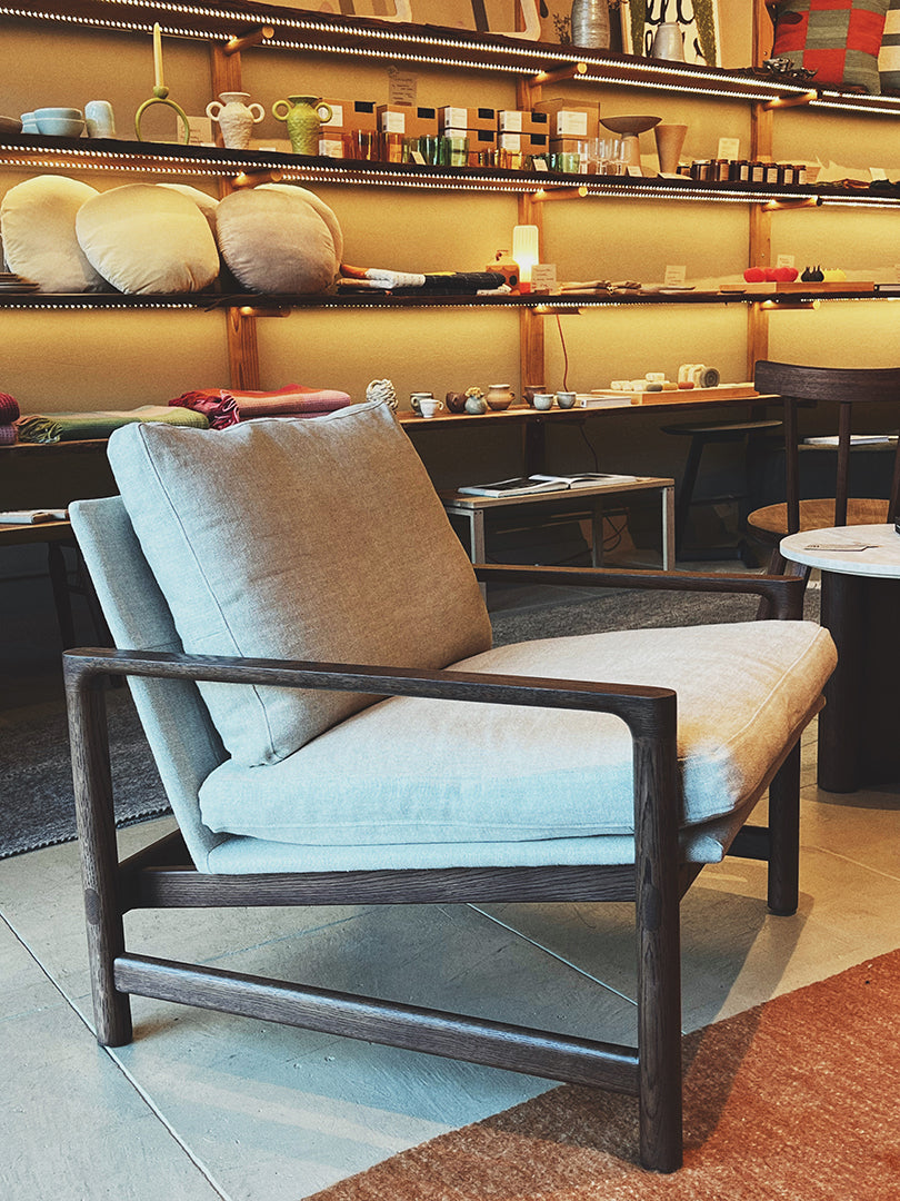 The Troubadour Lounge Chair | Fabric by Wooden Stories features light gray cushions, Danish lines, and a dark solid oak frame. Displayed in-store, it's surrounded by shelves of home decor, ceramics, and soft lighting.