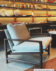 A Troubadour Lounge Chair | Fabric by Wooden Stories from the Danish Collection, with oatmeal-colored cushions and a solid oak frame in an espresso finish, sits in a cozy, well-lit store surrounded by shelves of home decor and ceramics. A small label reads, L653 Oatmeal with Espresso Oak.