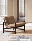 A Troubadour Lounge Chair | Fabric by Wooden Stories, featuring a solid oak frame and apricot-colored cushions, is placed on a light area rug in a sunlit room with large doors and patterned tile floors.