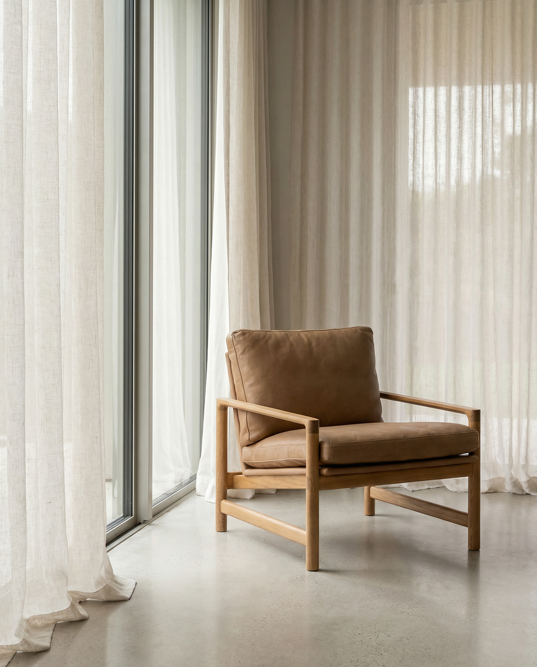 The Troubadour Lounge Chair | Leather by Wooden Stories, featuring a tan leather seat and solid oak frame, sits before tall windows with sheer beige curtains, crafting a calm, minimalist, sunlit space inspired by Danish design.