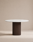 The Tathra Round Dining Table by Wooden Stories features a stone top and sturdy, cylindrical dark wood base, showcased against a neutral beige background.