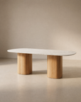 The Tathra Ellipse Dining Table by Wooden Stories features an oval stone top and two solid oak pedestal legs, highlighting European craftsmanship, set against a neutral beige background.
