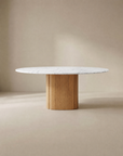 The Tathra Round Dining Table by Wooden Stories features a stone top and a cylindrical solid oak base, shown here with a white marble finish against a neutral beige background.