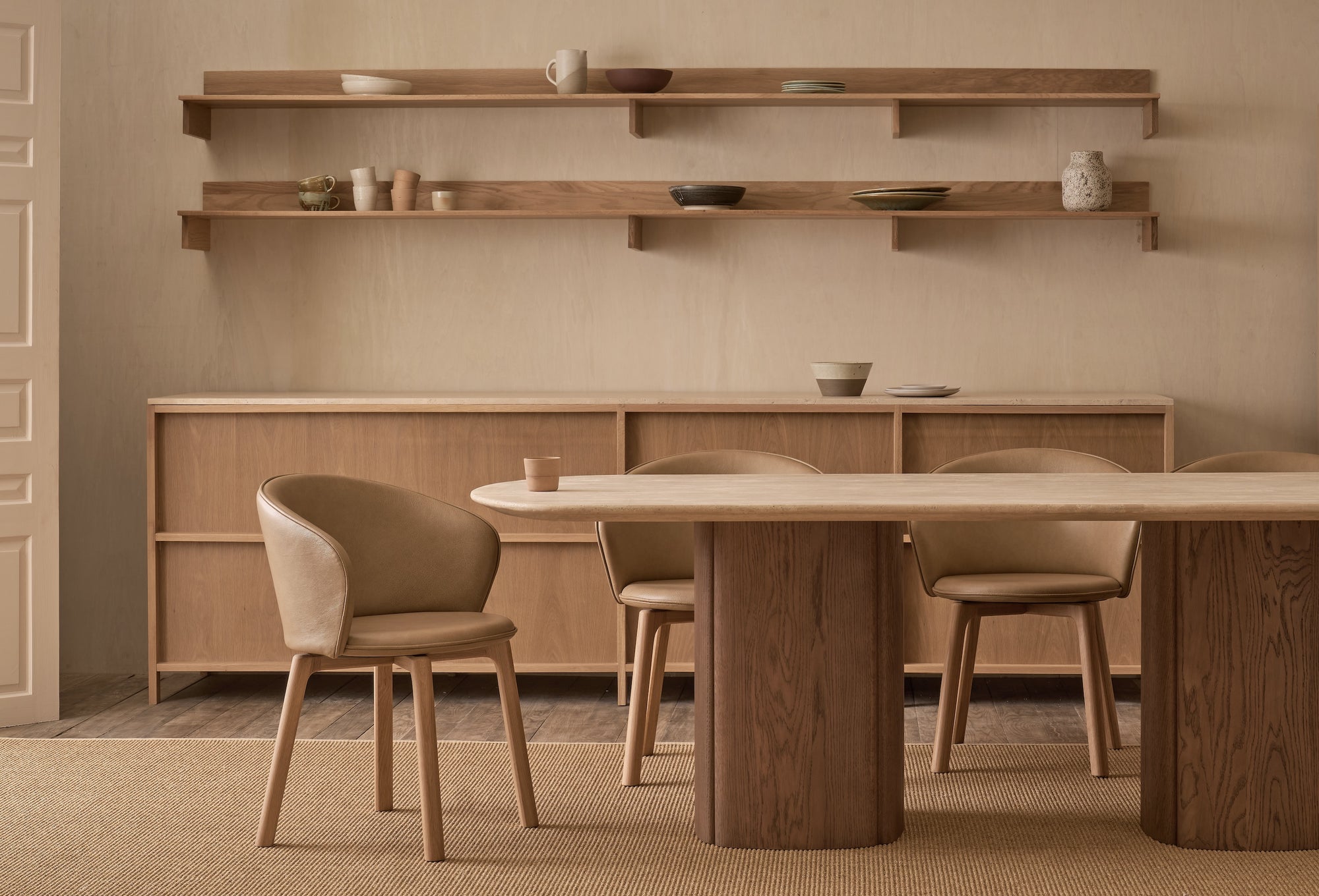 Minimalist dining room with light wood furniture, featuring the Wooden Stories Tathra Ellipse Dining Table | Stone Top, four upholstered chairs, a sideboard, shelves with ceramics, and a neutral palette that highlights natural textures.
