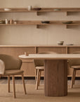 Minimalist dining room with light wood furniture, featuring the Wooden Stories Tathra Ellipse Dining Table | Stone Top, four upholstered chairs, a sideboard, shelves with ceramics, and a neutral palette that highlights natural textures.