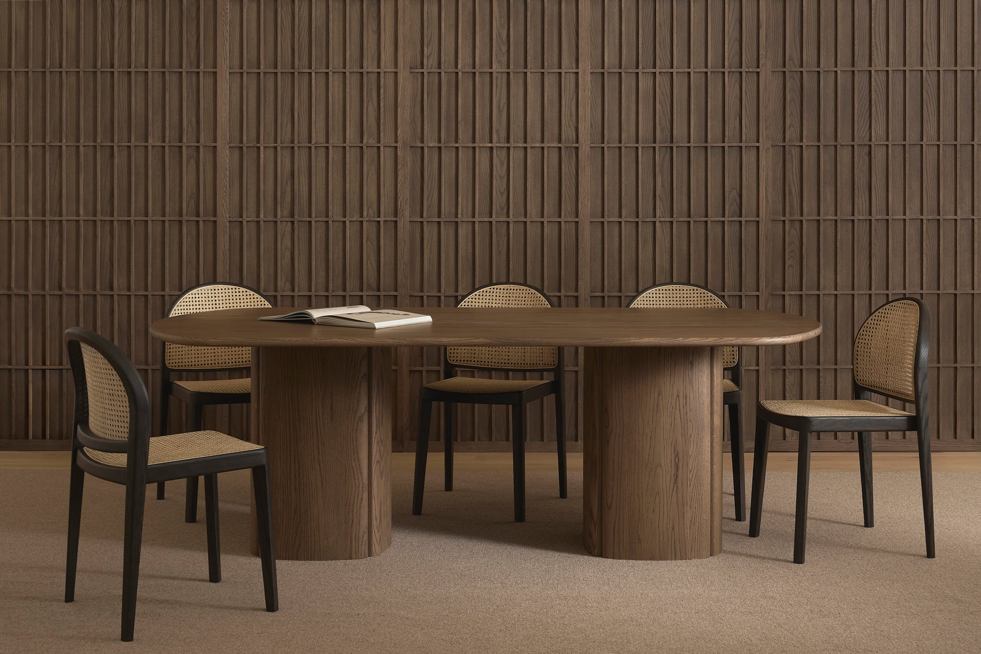 A modern dining room showcases the Wooden Stories Tathra Ellipse Dining Table with a stone top, accompanied by six black-framed chairs with woven cane backs, set against wood paneling and a beige carpet. An open book highlights European craftsmanship.
