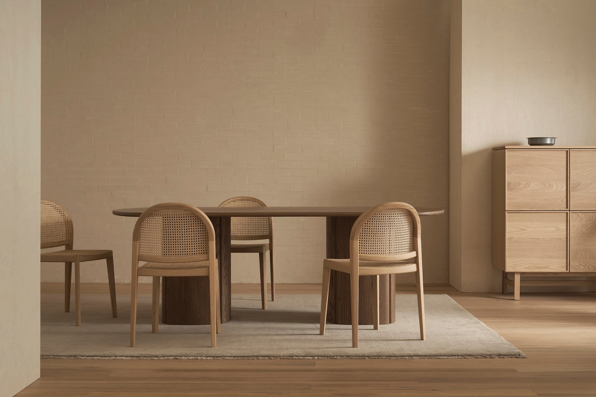 Minimalist dining room featuring the Tathra Ellipse Dining Table | Stone Top by Wooden Stories, four woven-back chairs, a light rug on wooden flooring, and a light wood cabinet with a small bowl, showcasing European craftsmanship.