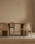 Minimalist dining room featuring the Tathra Ellipse Dining Table | Stone Top by Wooden Stories, four woven-back chairs, a light rug on wooden flooring, and a light wood cabinet with a small bowl, showcasing European craftsmanship.