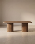 The Tathra Ellipse Dining Table | Oak by Wooden Stories features a modern solid oak oval top and two wide cylindrical legs, beautifully displayed in a minimalist room with neutral walls and flooring.