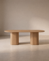 The Wooden Stories Tathra Ellipse Dining Table | Oak features a minimalist solid oak design with a smooth oval top and two cylindrical pedestal legs, elegantly situated in a softly lit, neutral-toned room.