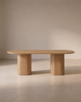 The Wooden Stories Tathra Ellipse Dining Table | Oak features a minimalist solid oak design with a smooth oval top and two cylindrical pedestal legs, elegantly situated in a softly lit, neutral-toned room.