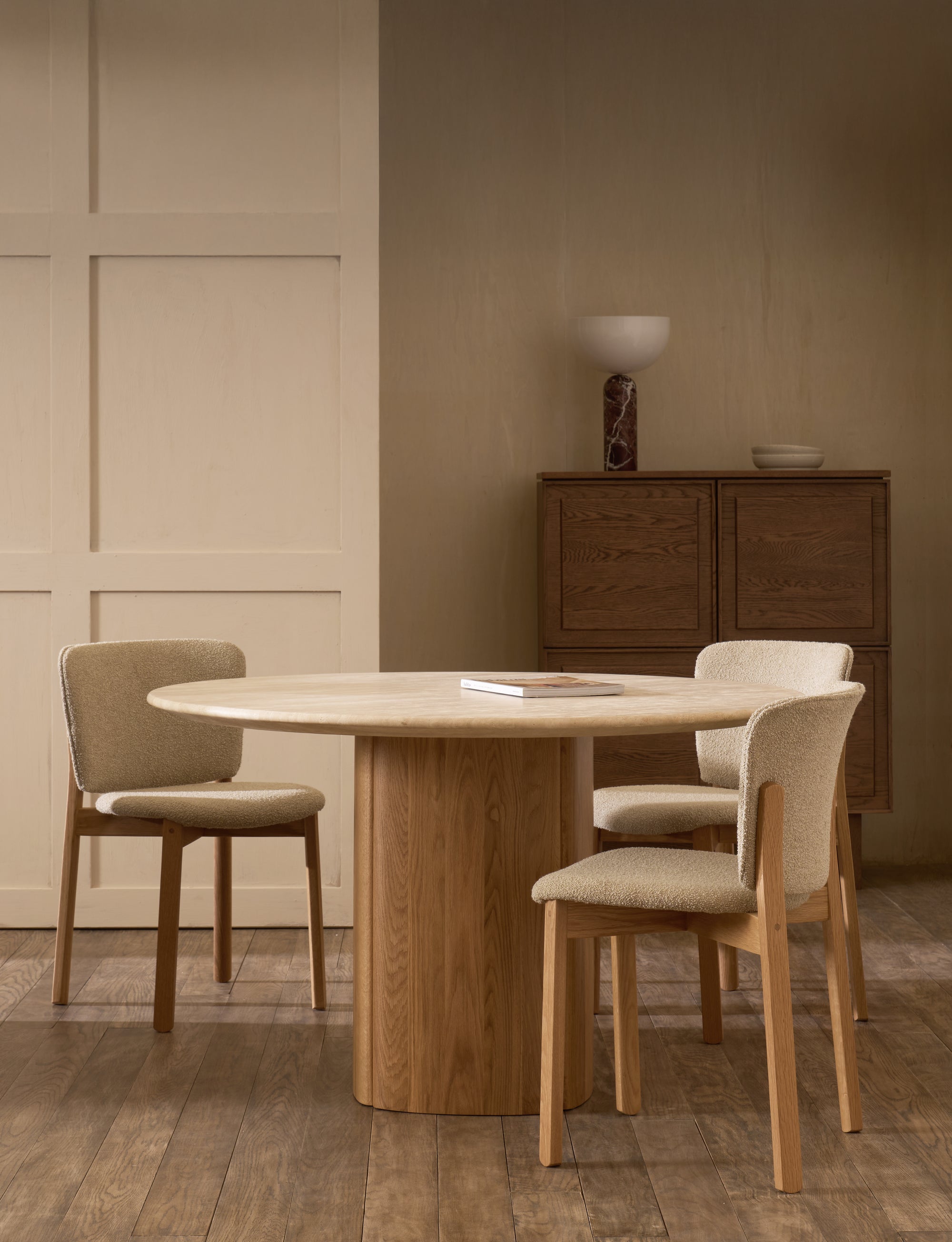 In a minimalist room, the Tathra Round Dining Table | Stone Top by Wooden Stories is paired with three upholstered chairs. A book rests on the contract-grade table, with a wooden cabinet, bowl, and lamp behind, all in neutral tones on wood flooring.