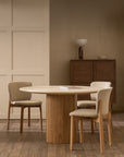 In a minimalist room, the Tathra Round Dining Table | Stone Top by Wooden Stories is paired with three upholstered chairs. A book rests on the contract-grade table, with a wooden cabinet, bowl, and lamp behind, all in neutral tones on wood flooring.