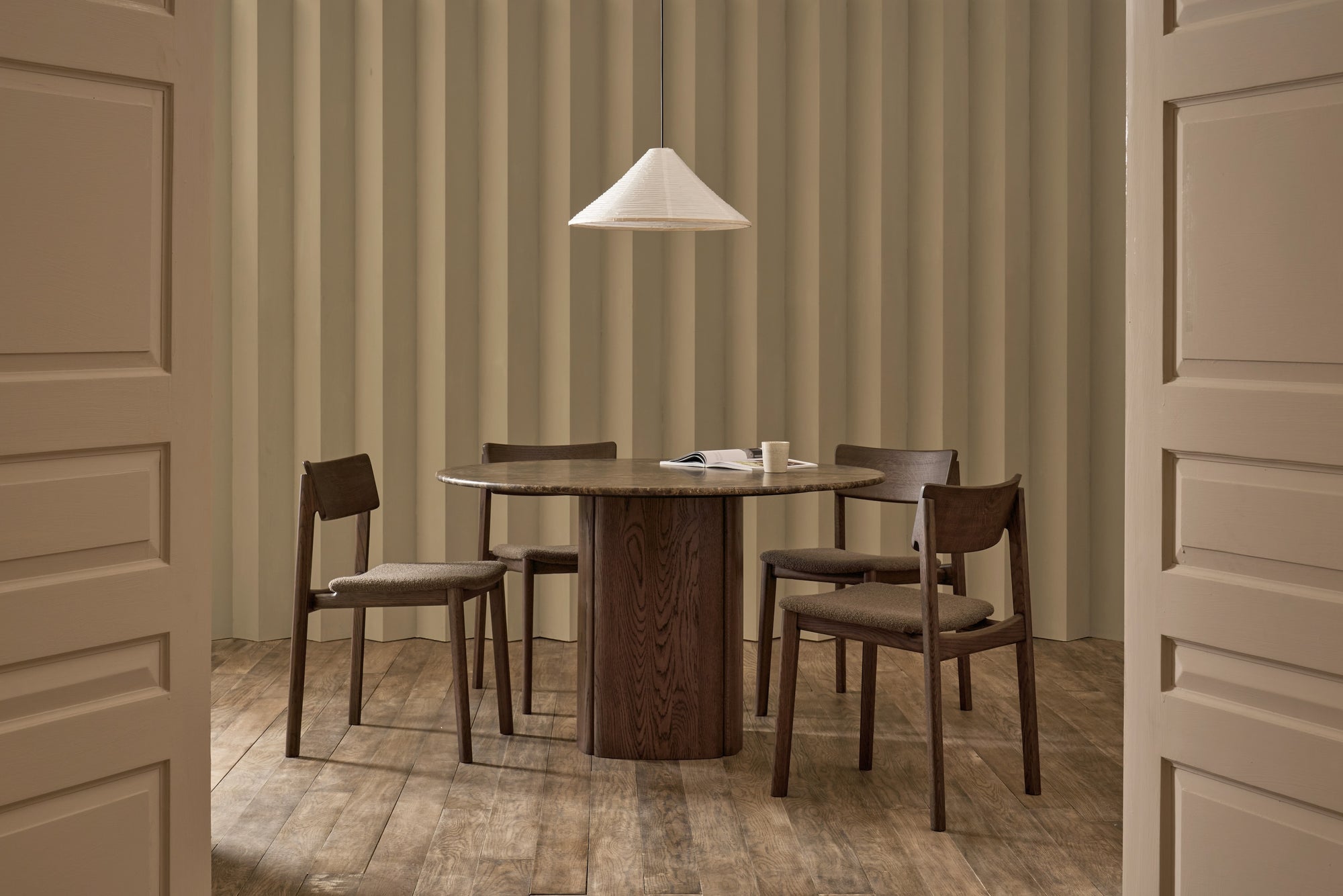 A minimalist dining room with the Wooden Stories Tathra Round Dining Table (stone top), four matching chairs, a white pendant lamp above, and a textured beige wall behind, viewed through open double doors. A cup and an open book rest on the table.