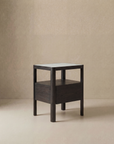 The Theo Nightstand by Wooden Stories is a minimalist dark wood side table with Danish lines, featuring a square top, four sturdy legs, and a lower shelf, shown on a light floor against a plain beige wall.