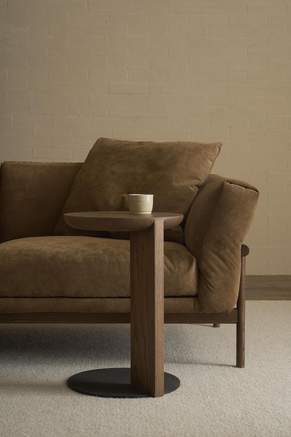 A brown cushioned sofa sits beside the Wooden Stories Guild Side Table in Espresso Oak, topped with a white cup, against a light beige brick wall and neutral carpeted floor.