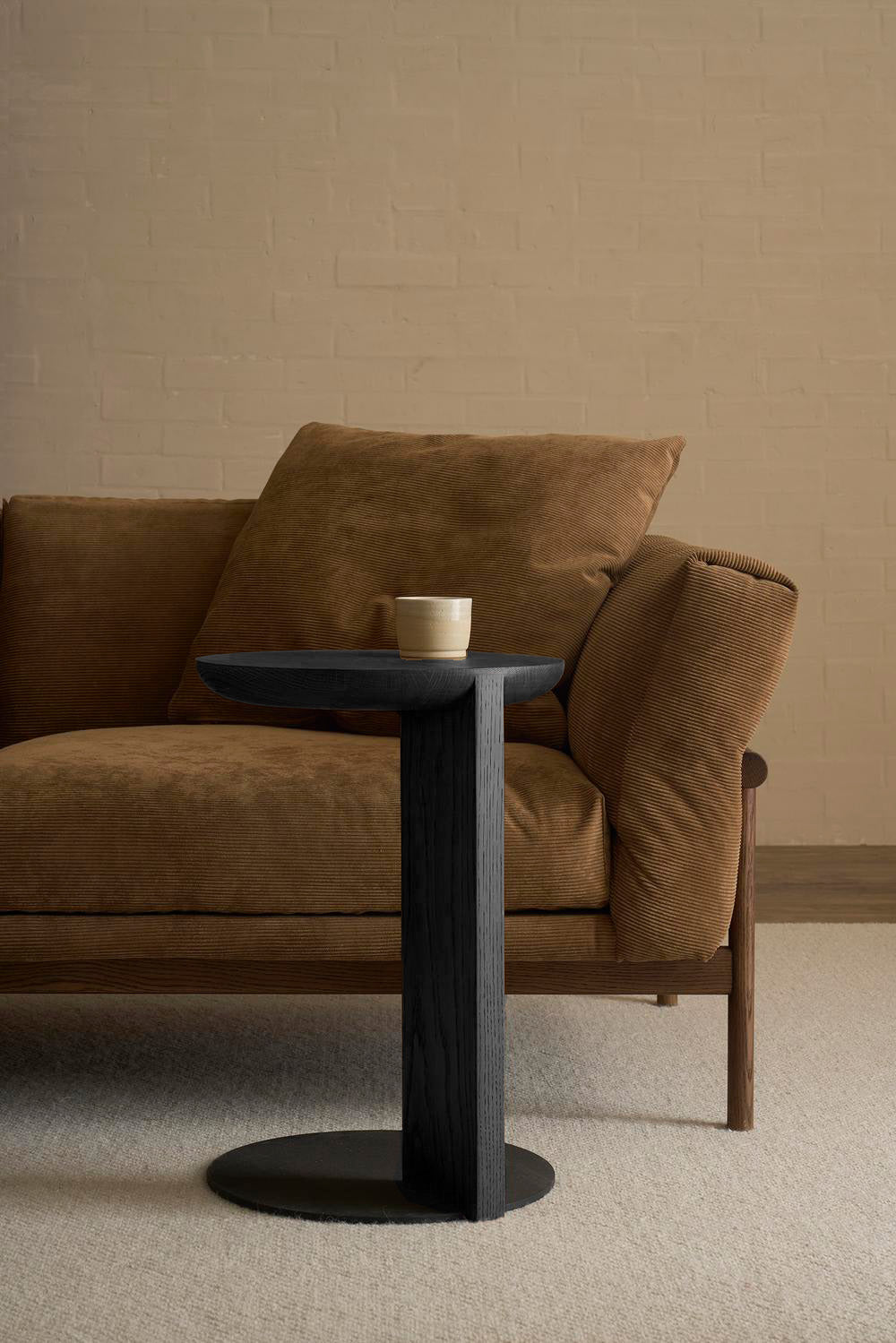 A modern brown velvet sofa with a wooden frame sits against a beige brick wall. In front, the Guild Side Table in black oak by Wooden Stories holds a single white cup, all on a light carpeted floor.
