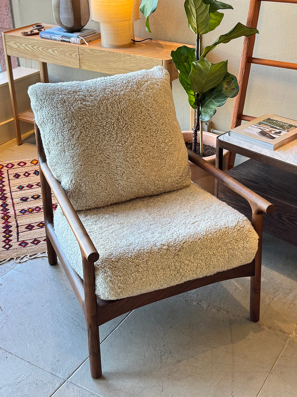 A SOBU Viking Lounge Chair - Walnut featuring a wooden frame and plush cream shearling cushions sits in a cozy living space beside a table, plant, patterned rug, and a small stack of books.
