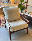 A SOBU Viking Lounge Chair - Walnut featuring a wooden frame and plush cream shearling cushions sits in a cozy living space beside a table, plant, patterned rug, and a small stack of books.