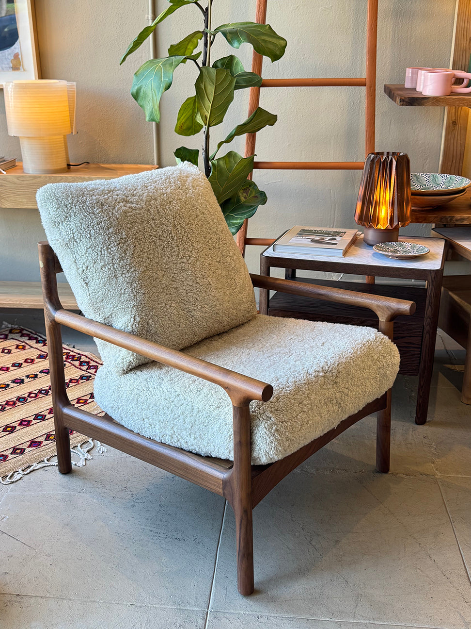 The SOBU Viking Lounge Chair - Walnut, featuring light, fuzzy upholstery and a dark wooden frame, sits beside a side table with a lamp, plant, dishes, and magazines in a modern living space.
