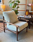 The SOBU Viking Lounge Chair - Walnut, featuring light, fuzzy upholstery and a dark wooden frame, sits beside a side table with a lamp, plant, dishes, and magazines in a modern living space.