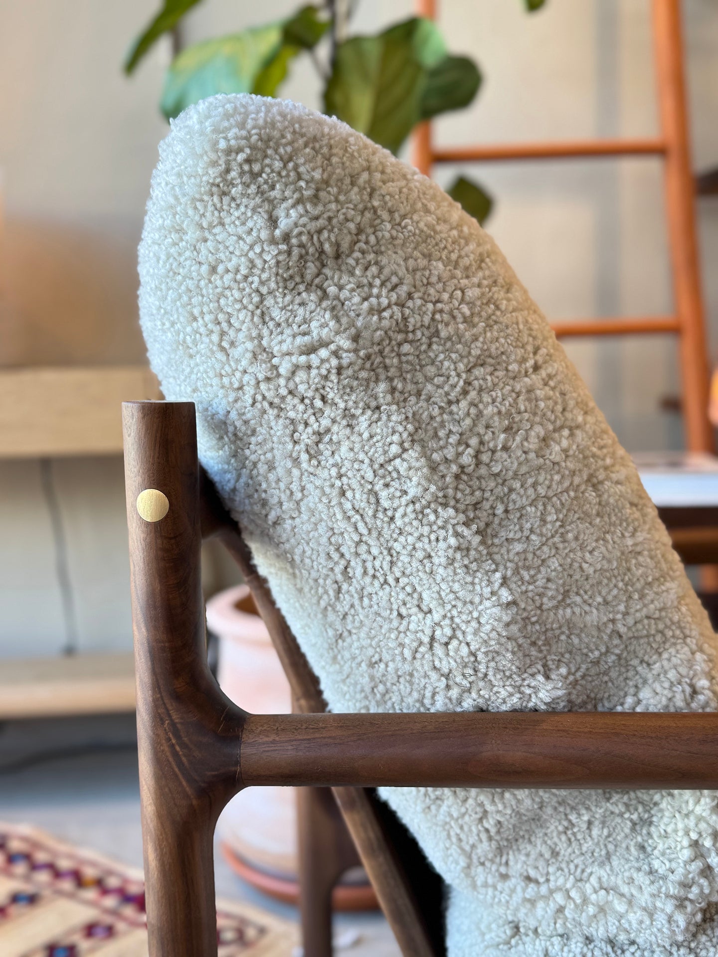 A close-up of SOBU’s Viking Lounge Chair – Walnut with a wooden frame and cream sherpa-style fabric. In the background: a potted plant, wooden ladder, and patterned rug showcase signature SOBU style.