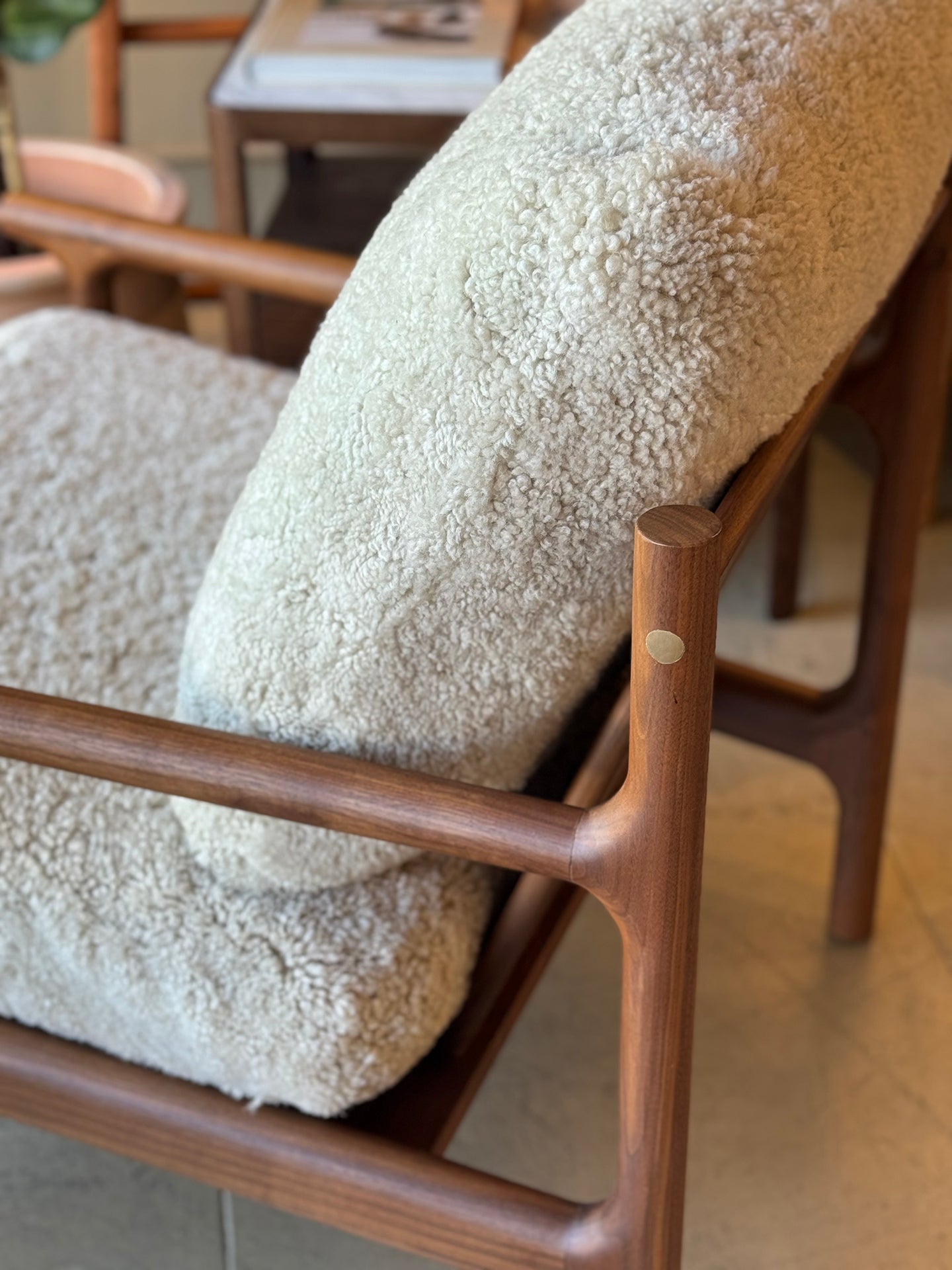 A close-up of the SOBU Viking Lounge Chair - Walnut, featuring solid walnut construction and soft, cream textured cushions, set in a warmly lit room with other SOBU furniture and wooden accents in the background.