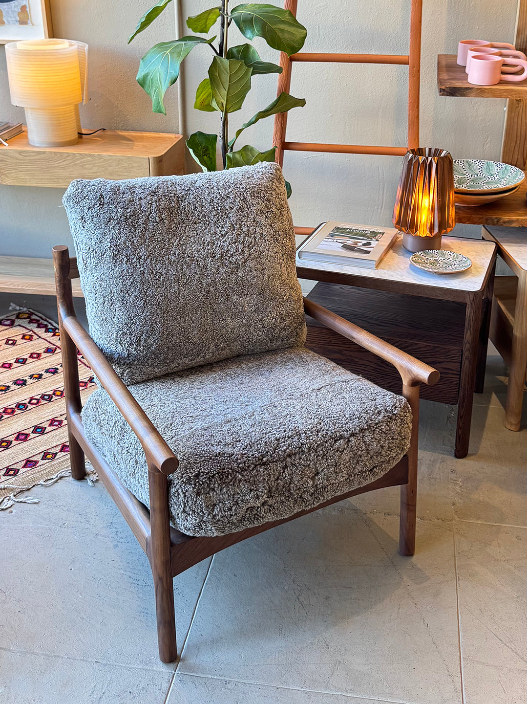 A stylish living space features the SOBU Viking Lounge Chair - Walnut with a gray textured cushion, placed next to a small table holding a lit lamp, books, and with a potted plant visible in the background.