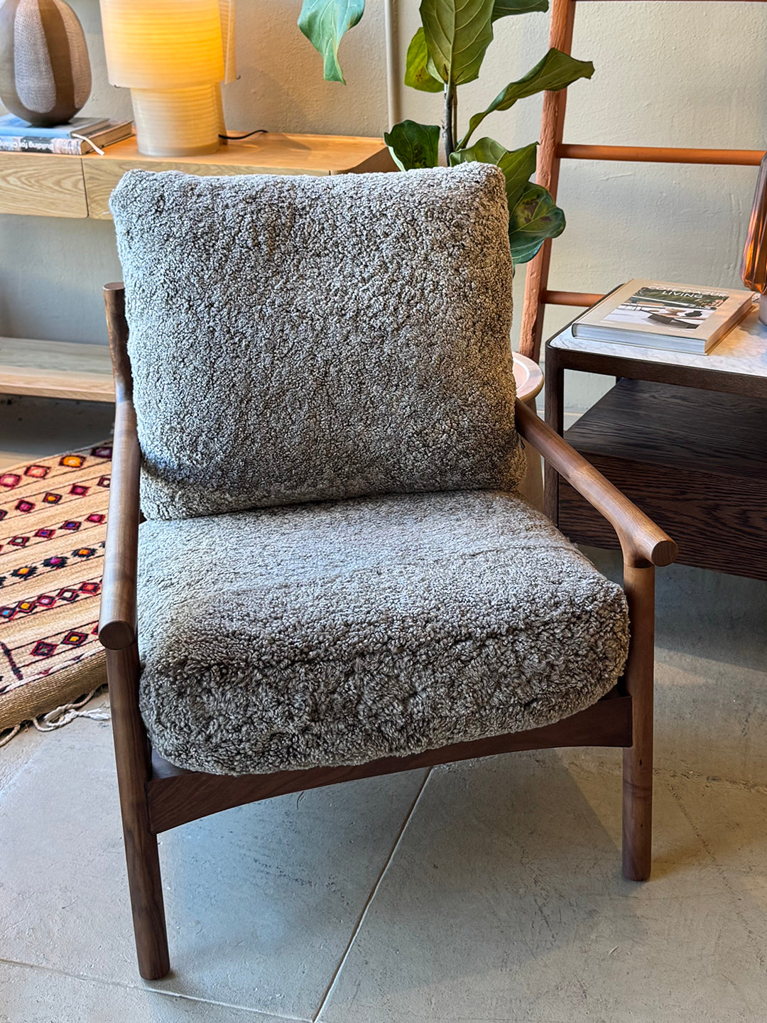 The Viking Lounge Chair - Walnut by SOBU, featuring plush textured gray cushions, sits beside a wooden table, plant, and lamp on a tiled floor—showcasing mid-century modern style in this cozy, well-lit room.