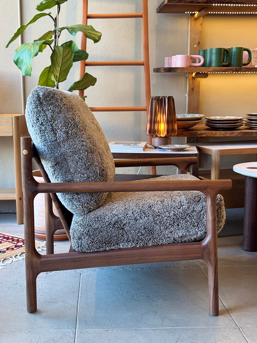 The Viking Lounge Chair - Walnut by SOBU, featuring plush textured gray cushions, sits beside a lamp-topped table. In the background, shelves with dishes and a potted plant highlight SOBU’s elegant furniture design.